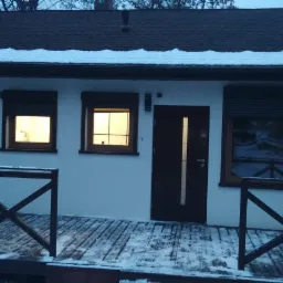 Dom jednorodzinny z drewnianym tarasem i częściowo opuszczonymi zewnętrznymi osłonami okiennymi w zimowej scenerii, widoczne numery domu '151'.